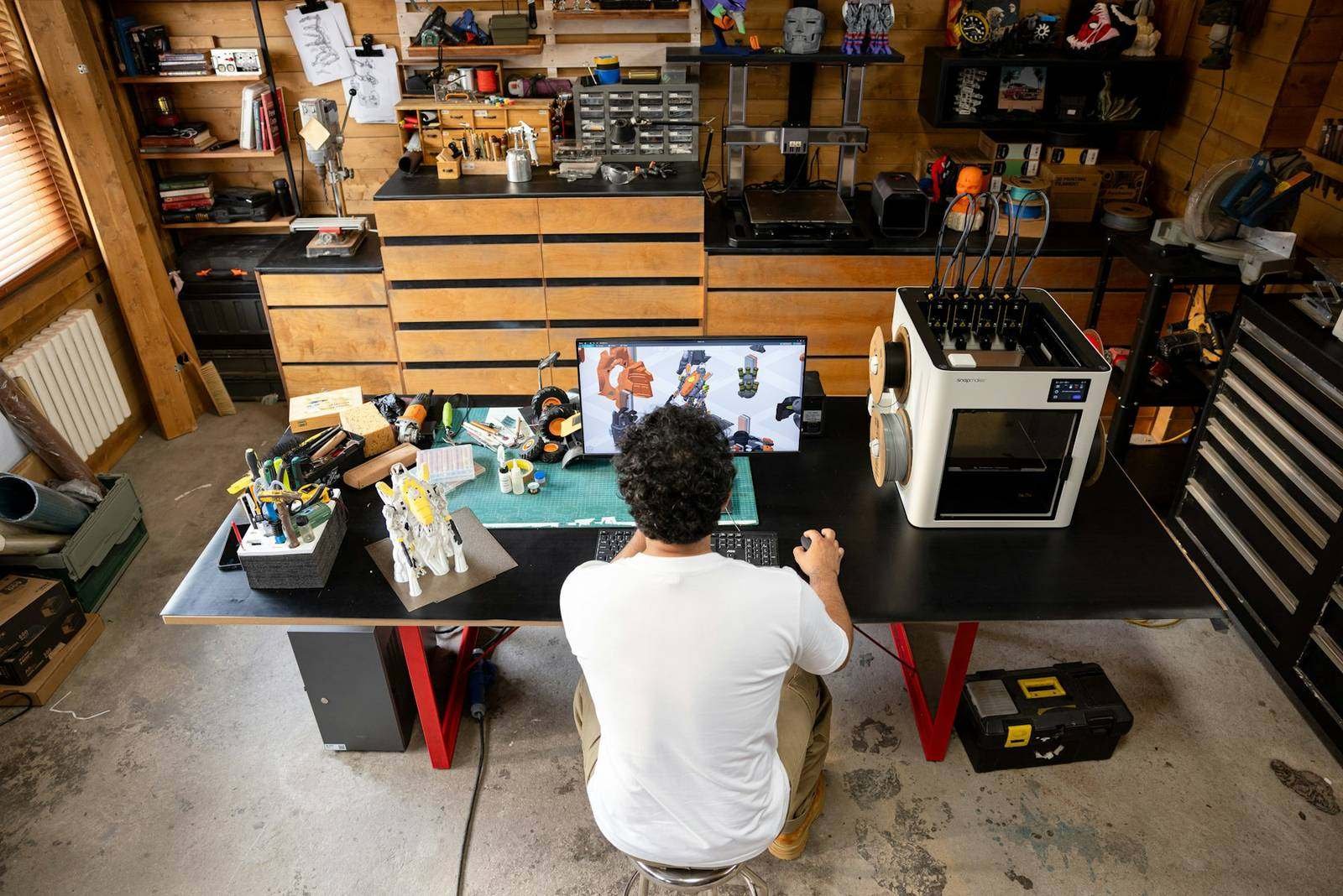 Designer at a studio workbench with a 3D printer, computer, hand tools and material drawers